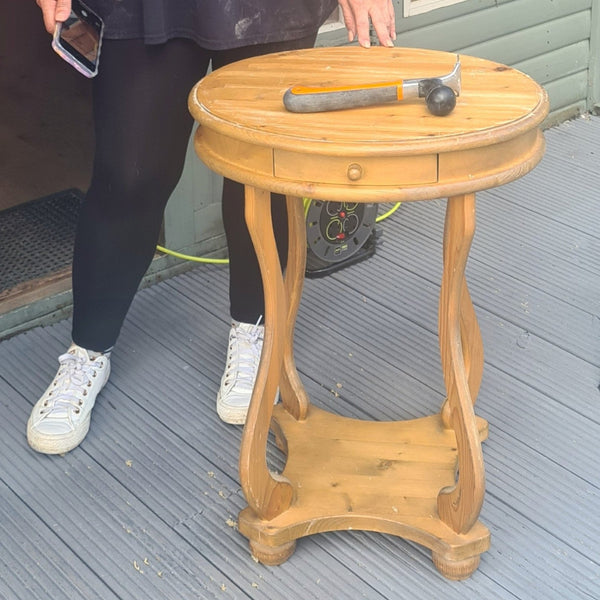 A pine wood round side table with a single drawer and curved legs, shown on a gray deck with a person standing beside, ready to start their workshop.
