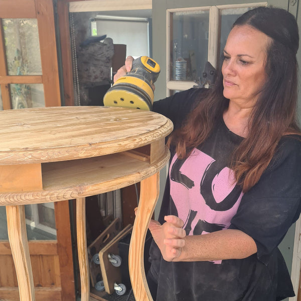 A woman in a black t-shirt holds a DeWalt random orbital sander against a wooden side table in a workshop setting.