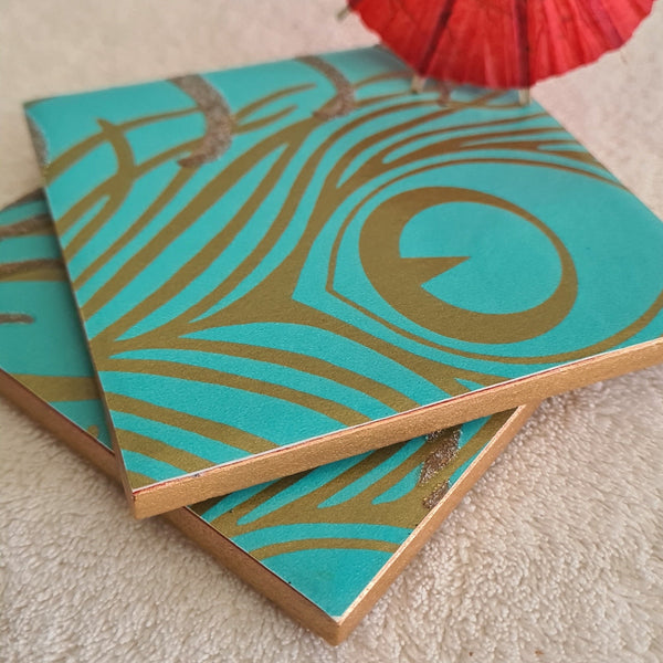 Two blue teal ceramic coasters with gold peacock feather design and added glitter.