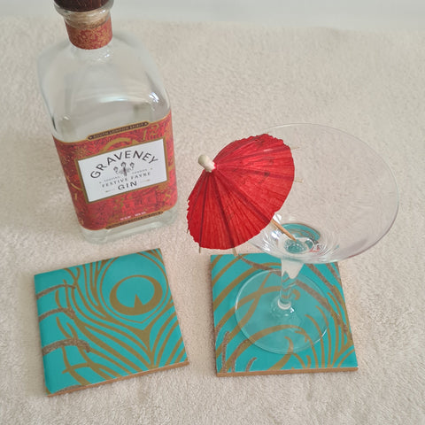 Gin bottle, cocktail glass and a red umbrella with two blue teal ceramic coasters with peacock feather pattern.