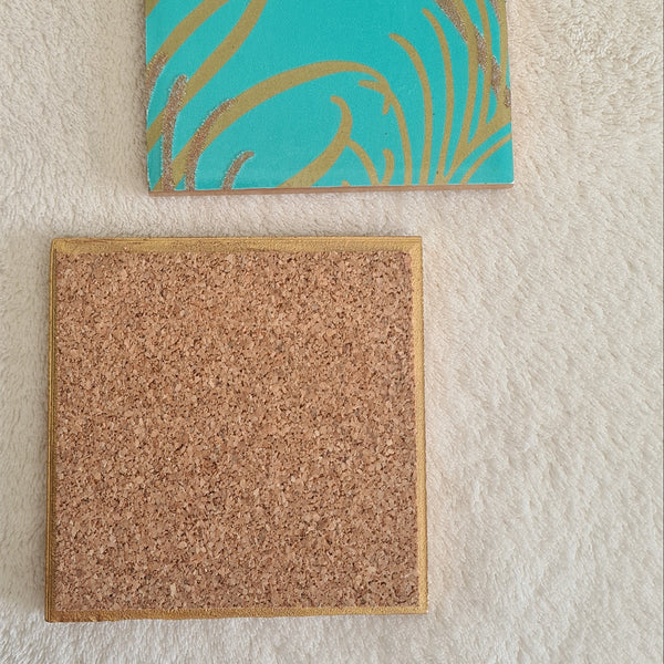 Two square ceramic coasters, one with a blue teal and gold peacock feather pattern, the other with a cork backing.