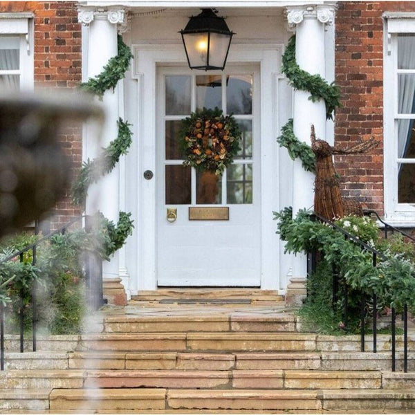 The white front door of Champneys, Henlow on a brick house porch decorated with green garlands, a floral wreath, stone steps, and a black lantern above the entrance.