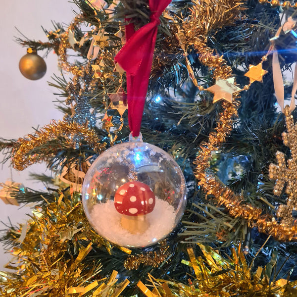 A handmade clear bauble snowglobe containing a handpainted wooden mushroom and snow, hanging on a Christmas tree with a red ribbon.