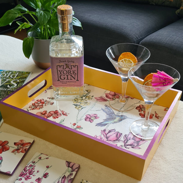 Deep yellow handcrafted wooden serving tray with integrated handles, hummingbird floral design and metallic violet edging, styled with cocktail glasses and a bottle.