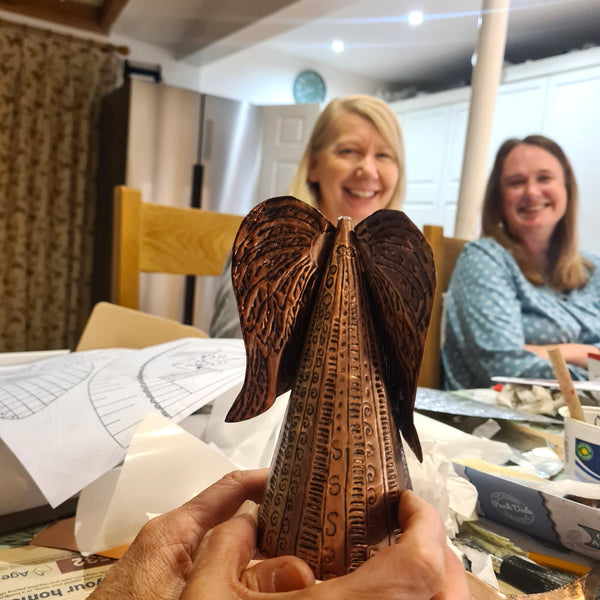 A beginner holding her metal embossed copper angel with detailed wings and script engravings, in a home workshop environment with two women smiling in the background.