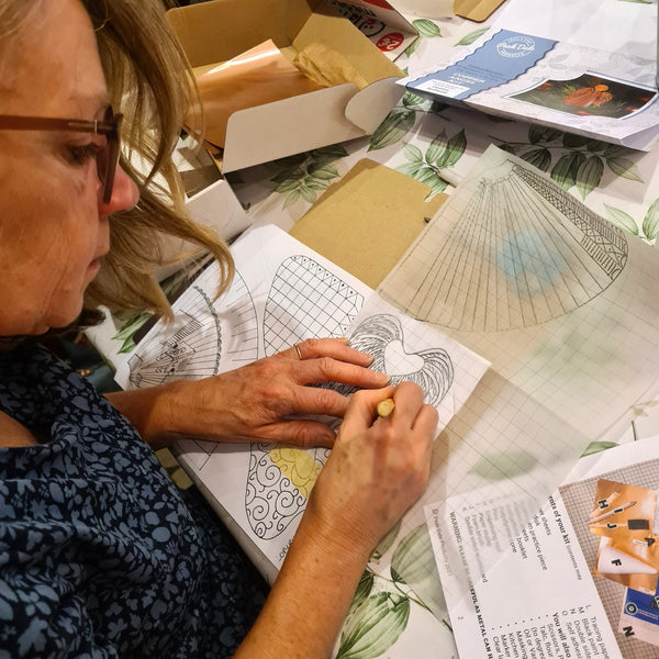A woman with glasses is carefully making her template, ready to emboss part of her copper angel, surrounded by craft supplies, papers, and boxes.