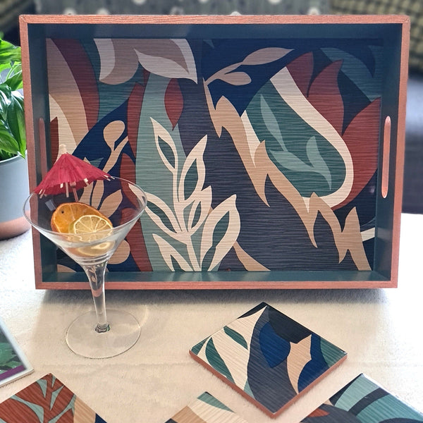 Handcrafted deep teal wooden serving tray decorated with a bold retro leaf design and metallic copper edging, styled on a coffee table with cocktail glass.