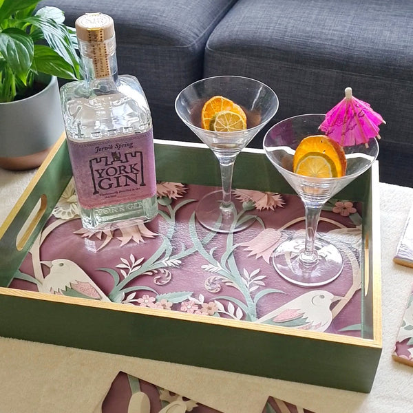 Handcrafted sage green wooden serving tray decorated with woodland animals and foliage on a rich plum background with metallic gold edging, styled on a coffee table.